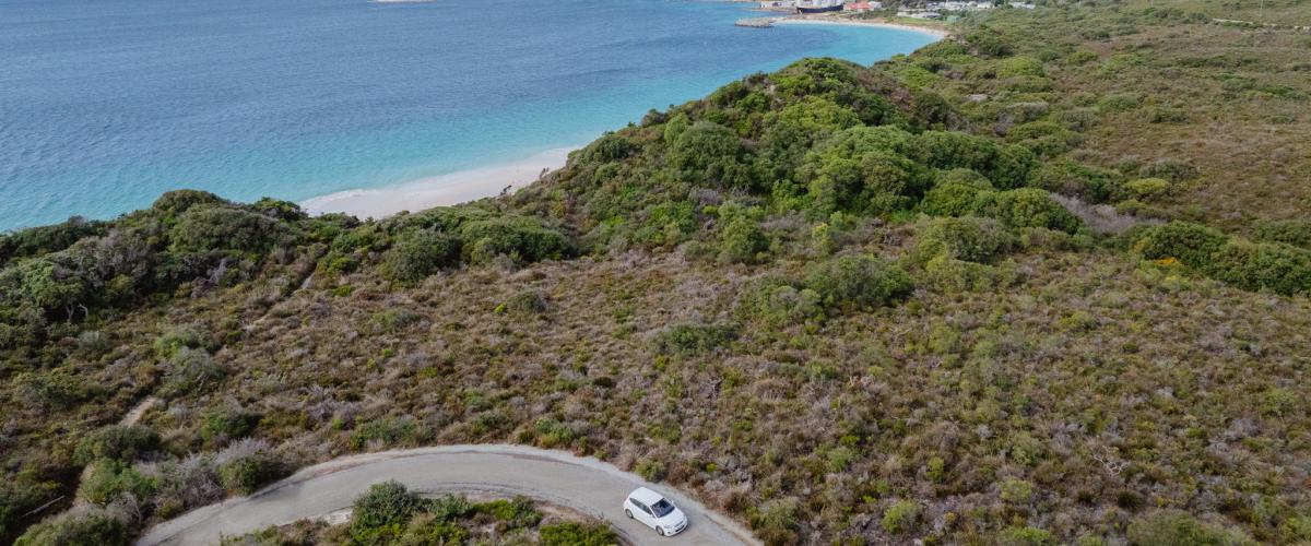 Aerial view of a car on a road winding through bushland with a beach and ocean in the distance